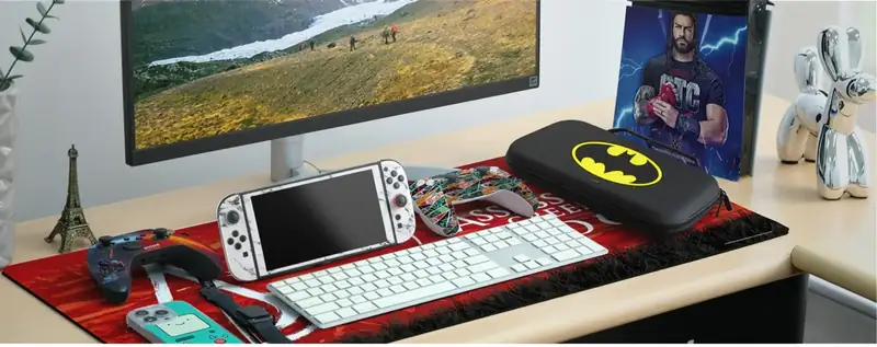 A gaming desk with an Assassin's Creed desk mat, a Batman Nintendo Switch carry case, a skinned Switch OLED and skinned controllers